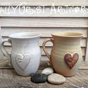 May include: Two ceramic mugs, one off-white and one tan, each featuring a heart with a paw print. They have large handles and sit on a wooden surface. The text "www.SallyGrist-Artwork.co.uk" is visible at the top.