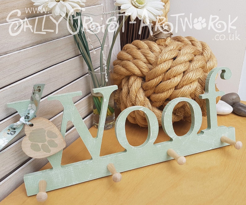 May include: A light green wooden wall hook with the word "Woof" cut out. The hook has three pegs and a heart-shaped tag with a paw print design. A ribbon with paw prints is attached to the tag. The background includes a decorative rope knot and flowers.