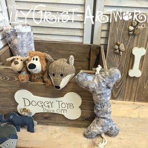 May include: A dark brown wooden storage crate featuring a cream-colored bone-shaped plaque with the text "Doggy Toys Paws Off!" and a matching lid. The crate contains plush animal toys, and a bone-shaped fabric toy with a paw print pattern sits in front.