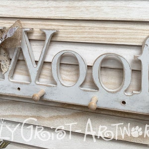 May include: A distressed white wooden wall-mounted coat rack with the word "WOOF" in large letters. It has four wooden pegs for hanging items and is decorated with a paw print heart and a burlap bow.