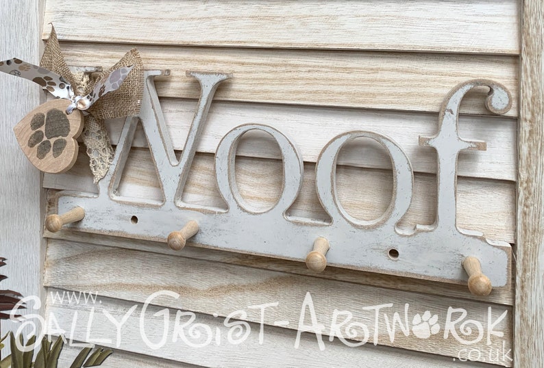 May include: A distressed white wooden wall-mounted coat rack with the word "WOOF" in large letters. It has four wooden pegs for hanging items and is decorated with a paw print heart and a burlap bow.