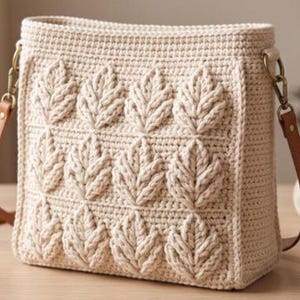 May include: A cream-colored crochet shoulder bag featuring a textured leaf pattern on the front panel. The bag includes a brown leather strap attached with antique brass-toned metal hardware clips on both sides of the rectangular body.