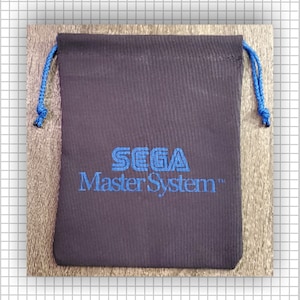 May include: A black fabric drawstring pouch featuring the blue embroidered text SEGA Master System centered on the front. The pouch includes two blue braided cords at the top for closure, providing a secure way to store small items or gaming accessories.