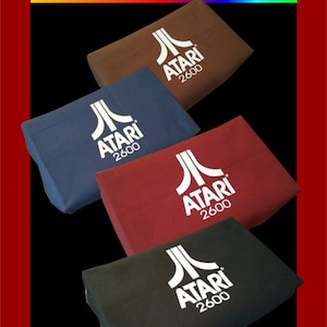 May include: Four Atari 2600 dust covers in brown, navy blue, burgundy, and black. Each cover has the Atari logo and "2600" printed in white. The covers are rectangular and angled, with a black background and a red border.