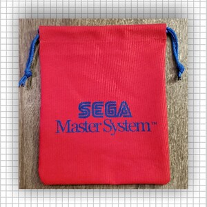 May include: A red fabric drawstring pouch featuring the blue embroidered text SEGA Master System centered on the front. The bag includes blue cord drawstrings on both sides, providing a secure closure for the rectangular storage accessory.