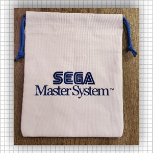 May include: A white fabric drawstring pouch featuring the blue SEGA Master System logo printed on the front. The bag includes blue cord drawstrings on both sides, tied with knots at the ends, providing a secure closure for the rectangular pouch.