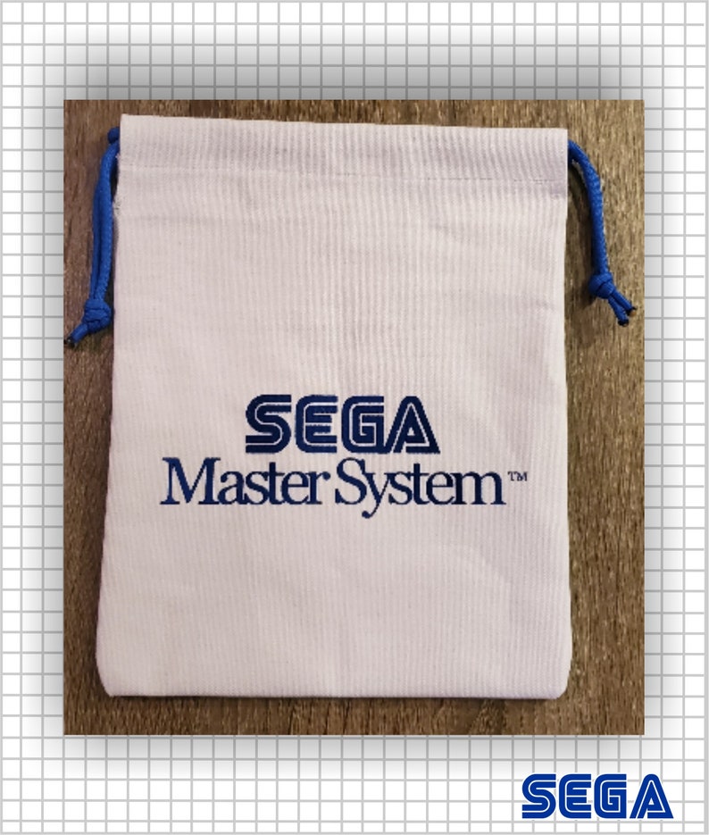 May include: A white fabric drawstring pouch featuring the blue SEGA Master System logo printed on the front. The bag includes blue cord drawstrings on both sides, tied with knots at the ends, providing a secure closure for the rectangular pouch.