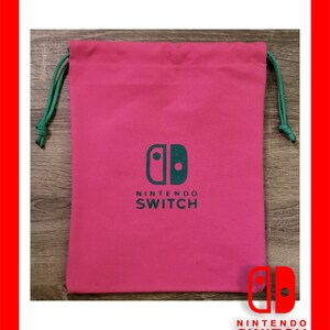 May include: A pink drawstring bag with a green drawstring. The bag features a green Nintendo Switch logo and the words "NINTENDO SWITCH" printed below. The bag is rectangular and made of a soft material.