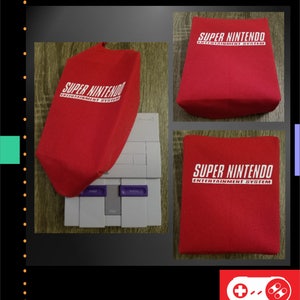 Op de afbeelding: Rode stoffen hoes met de tekst "SUPER NINTENDO ENTERTAINMENT SYSTEM" in het wit. De hoes wordt in meerdere weergaven getoond, waaronder een die een grijze Super Nintendo console bedekt. De afbeelding bevat ook een "CLASSICS" logo.