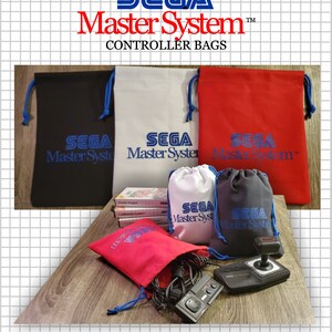 May include: A collection of drawstring bags in black, white, and red, each featuring the blue Sega Master System logo. The set includes large flat bags and smaller pouches, with some shown containing black video game controllers and game cartridges.