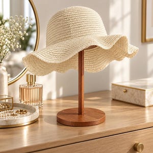 May include: A cream-coloured woven straw hat with a wide, wavy brim resting on a dark brown wooden stand. The hat features a rounded crown and a textured, open-weave construction, displayed on a circular base with a vertical support rod.
