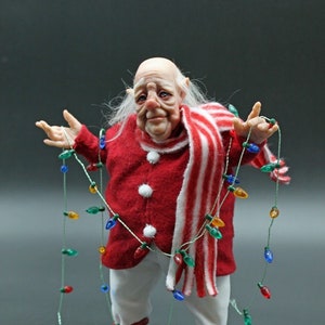 Puede incluir: Un duende de Navidad caprichoso con cabello blanco y una bufanda a rayas rojas y blancas, sosteniendo una cadena de luces navideñas de colores.