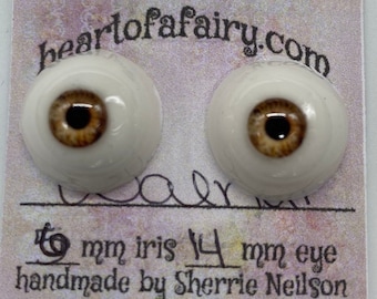 Puede incluir: Dos ojos de muñeca de vidrio de 14 mm con iris marrones y pupilas negras. Los iris tienen un anillo exterior marrón claro y un anillo interior marrón oscuro. Los ojos están en una tarjeta blanca con el texto "heartof afairy.com" y "12 mm iris 14 mm eye handmade by Sherrie Neilson".