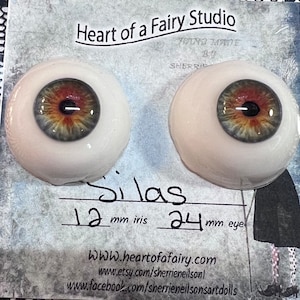 May include: A pair of realistic doll eyes with a green, brown, and orange iris. The eyes are 24 mm in diameter and the iris is 12 mm. The eyes are labelled "Silas".