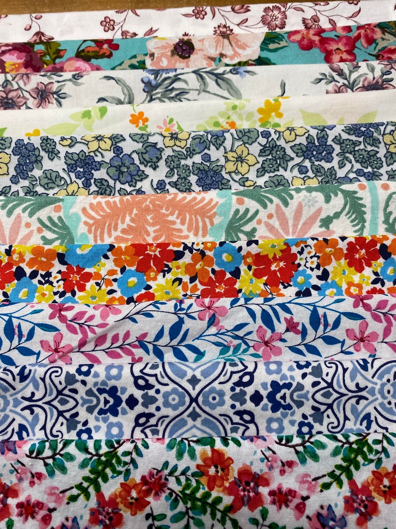 May include: A collection of fabric strips with various floral patterns in shades of pink, blue, green, and red. The fabric is arranged in a stack, with the top strip being a white background with a pink floral pattern.