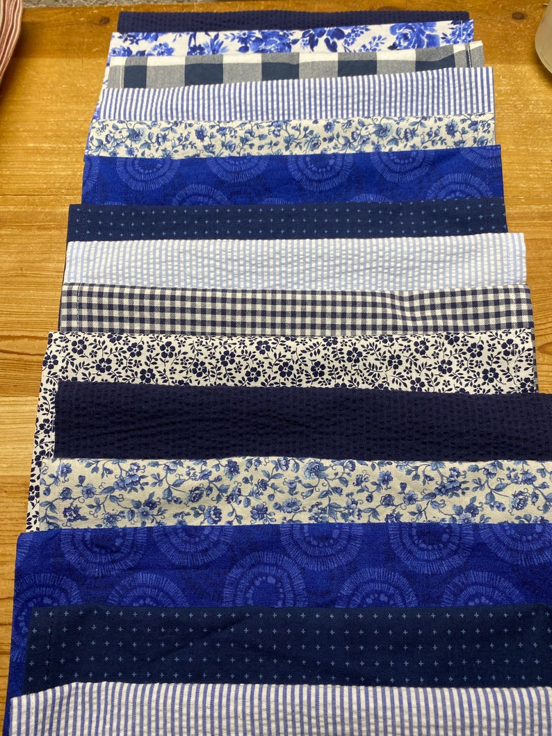 May include: A stack of 14 blue and white fabric squares with various patterns, including floral, gingham, and stripes. The fabric squares are arranged in a vertical stack, with the top square being a blue and white floral pattern.