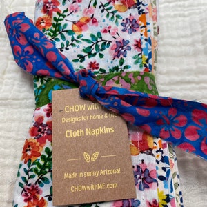 May include: A set of floral cloth napkins tied with a blue and pink fabric ribbon. The napkins are made in Arizona and feature a variety of colorful flowers on a white background. The tag reads "CHOW with Designs for home & life Cloth Napkins Made in sunny Arizona! CHOWwithME.com"