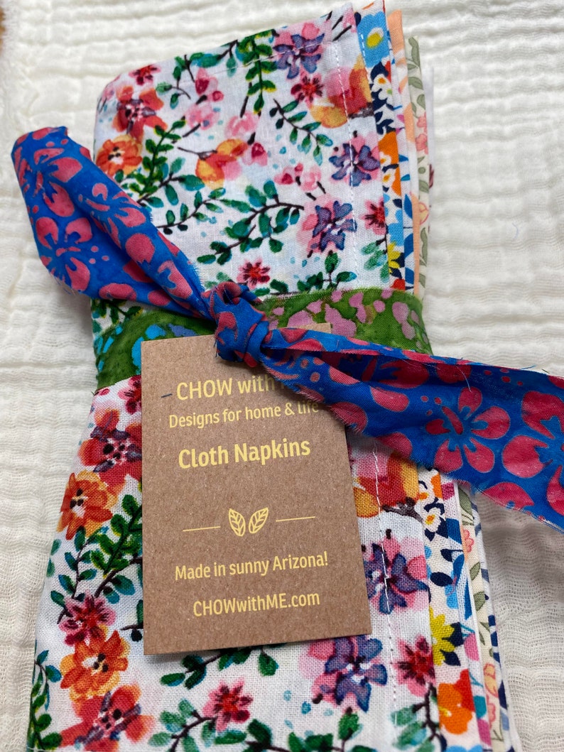 May include: A set of floral cloth napkins tied with a blue and pink fabric ribbon. The napkins are made in Arizona and feature a variety of colorful flowers on a white background. The tag reads "CHOW with Designs for home & life Cloth Napkins Made in sunny Arizona! CHOWwithME.com"