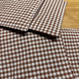 May include: Close-up of brown and white gingham fabric coasters. The coasters are square and have a classic checkered pattern. The fabric is tightly woven, and the edges are neatly sewn. The coasters are stacked on top of each other.