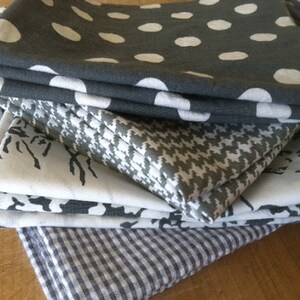 May include: A stack of six gray and white patterned fabric napkins. The top napkin has a white polka dot pattern on a gray background. The second napkin has a white and gray houndstooth pattern. The third and fourth napkins have a white floral pattern on a gray background. The fifth napkin has a white and gray geometric pattern. The bottom napkin has a white and gray gingham pattern.