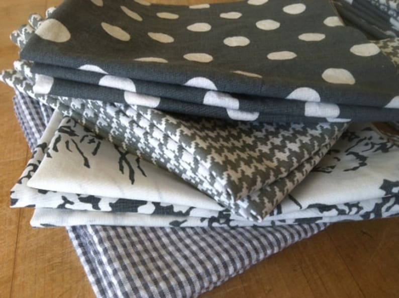 May include: A stack of six gray and white patterned fabric napkins. The top napkin has a white polka dot pattern on a gray background. The second napkin has a white and gray houndstooth pattern. The third and fourth napkins have a white floral pattern on a gray background. The fifth napkin has a white and gray geometric pattern. The bottom napkin has a white and gray gingham pattern.