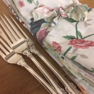 May include: Three silver forks with ornate handles, lying on a wooden surface. A stack of floral patterned napkins is tied with a ribbon and placed behind the forks.