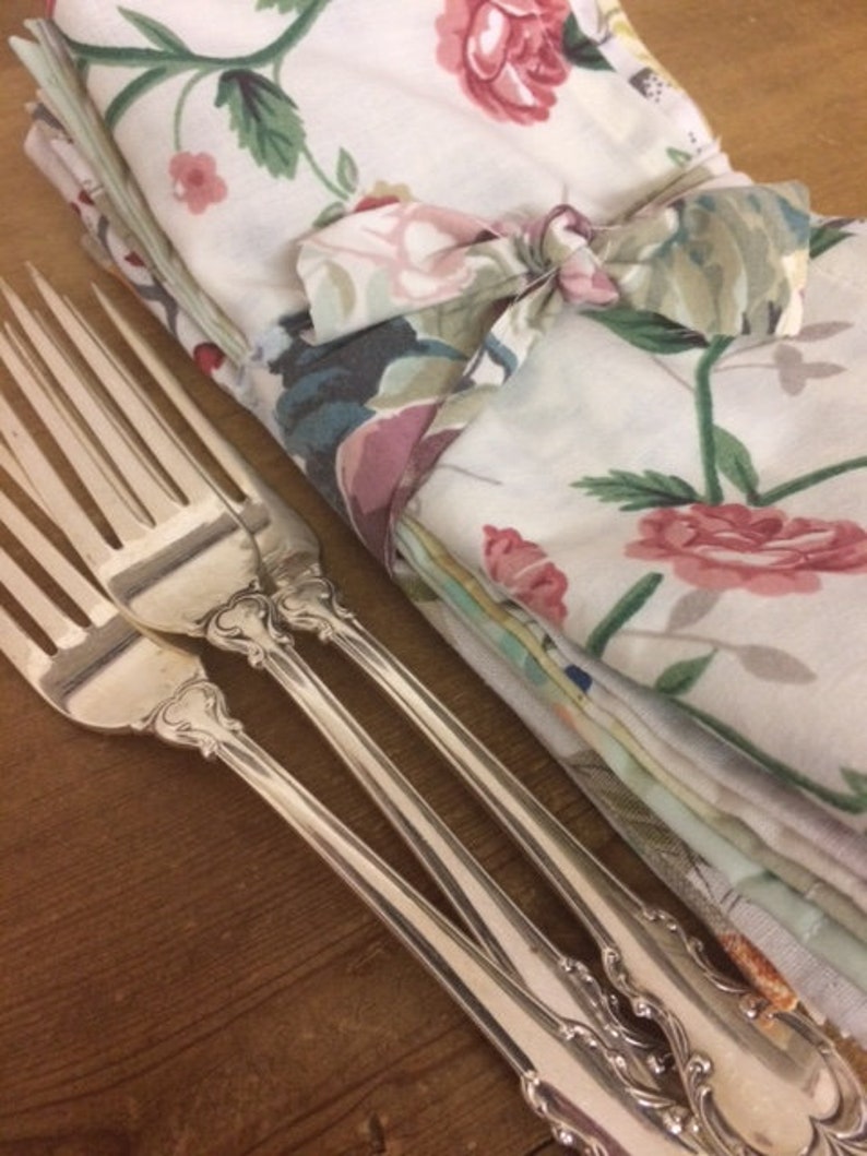 May include: Three silver forks with ornate handles, lying on a wooden surface. A stack of floral patterned napkins is tied with a ribbon and placed behind the forks.