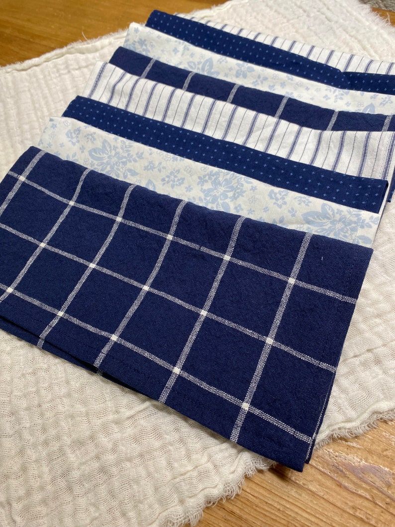 May include: A stack of seven blue and white patterned fabric napkins. The napkins are folded and arranged in a neat pile. The top napkin has a white grid pattern on a dark blue background.