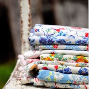 May include: A stack of vintage floral patterned fabric napkins in shades of blue, white, and red. The napkins are folded and stacked on a white wooden surface.
