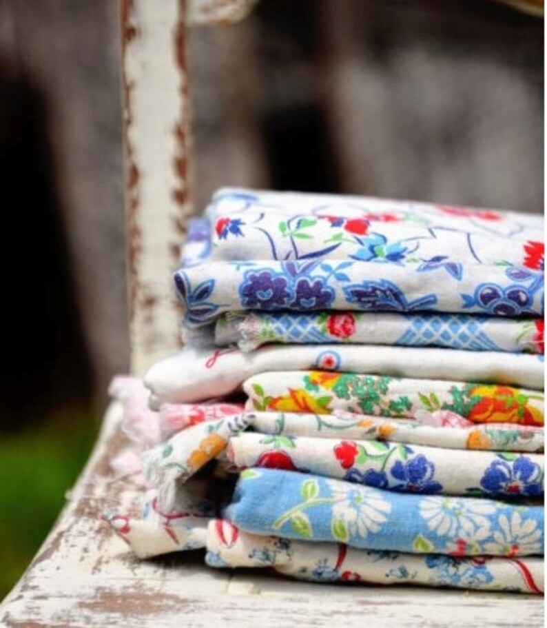 May include: A stack of vintage floral patterned fabric napkins in shades of blue, white, and red. The napkins are folded and stacked on a white wooden surface.