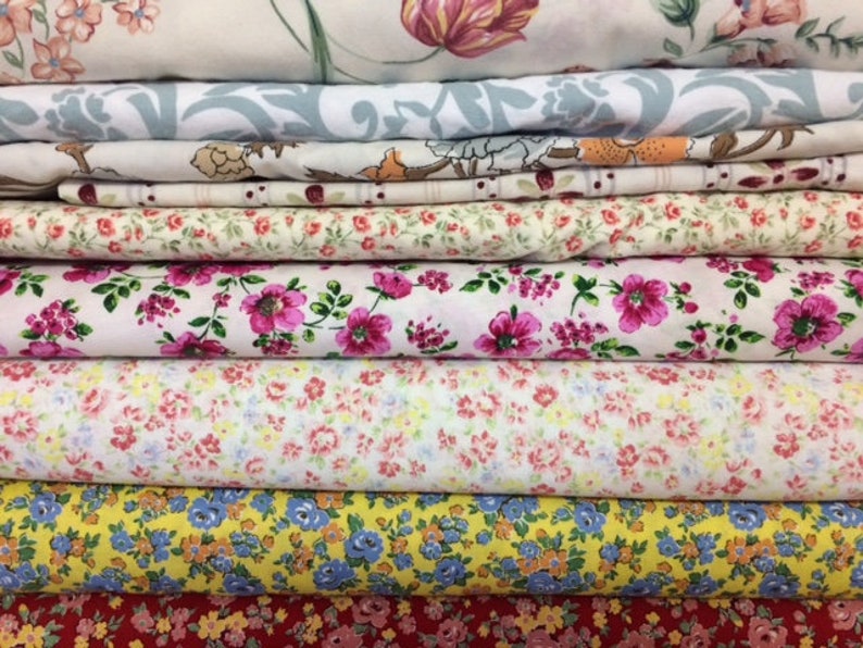 May include: A stack of eight fabric rolls with various floral patterns. The fabrics are in shades of pink, yellow, red, and blue.