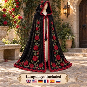May include: A black hooded cloak featuring intricate red rose appliqués with green stems and leaves. The garment has a scalloped red crochet trim along the hood and front edges, with a long black cord tie closure featuring decorative tassels at the ends.