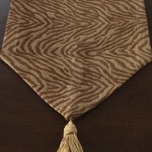 May include: A brown and beige table runner with a zebra print pattern and gold tassels on each end. The runner is laid out on a dark brown wooden table.