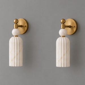 May include: A pair of wall sconces featuring a circular brushed brass backplate, a small spherical white marble accent, and a cylindrical, vertically fluted white marble shade. The fixtures are mounted vertically with a brass arm.