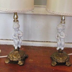 May include: Pair of white porcelain cherub table lamps with gold bases and white fabric lampshades with a white trim.
