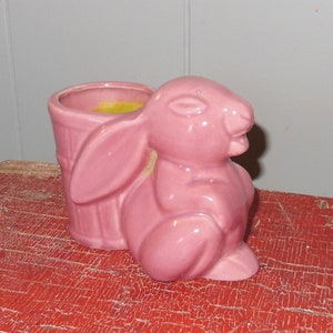 May include: A pink ceramic planter shaped like a rabbit. The planter has a small opening at the top for planting. The planter is sitting on a red wooden surface.