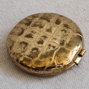 May include: A vintage, round, gold-toned compact with a textured, scale-like pattern on the lid. The compact has a small clasp and is slightly tarnished, giving it a classic, antique appearance. The compact is a beauty accessory.