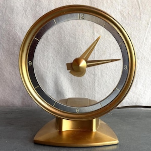 May include: A gold-toned, mid-century modern desk clock. The clock features a round face with gold hands and a gold bezel. The base is a matching gold color. The clock is powered by an electrical cord.