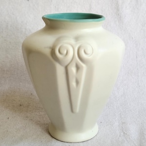 May include: Cream-colored ceramic vase with a teal interior rim. The vase has a geometric, faceted shape and a decorative embossed design on the front. The design includes two swirling shapes at the top and a diamond shape below.