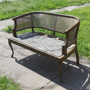 May include: A brown wooden bench with a woven cane back and seat. The bench has a dark gray cushion and sits on a concrete patio.