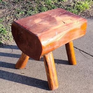 May include: A rustic wooden stool with a natural wood finish and three legs. The stool is made from a single piece of wood and has a rounded top.