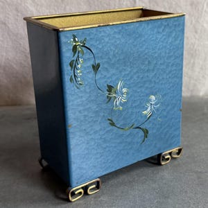 May include: A rectangular blue metal container with a hammered texture and a gold-toned rim. The front features a hand-painted floral design with white and green vines. The container is supported by four small, decorative gold-toned metal feet.