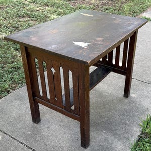 May include: A rectangular dark brown wooden side table featuring a flat top with visible surface wear and white markings. The base includes four straight legs connected by vertical slats with a diamond-shaped cutout and a lower horizontal shelf.