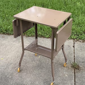 May include: A vintage brown metal side table with drop-leaf extensions. The table has a rectangular top, a lower shelf, and four legs with yellow caster wheels. The table is approximately 24 inches tall.