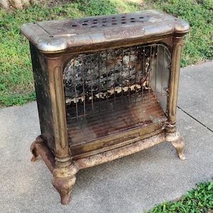 May include: An antique, cast iron heater with a rectangular top and ornate legs. The heater has a metal grate and a rusted, aged appearance. The top has several ventilation holes.