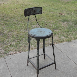 May include: A vintage metal stool with a green painted finish and a backrest. The stool has a round seat and a simple, sturdy frame.