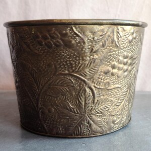 May include: A vintage metal planter with an embossed grape and leaf design. The planter has a round shape with a slightly flared rim. The metal has a tarnished, antique appearance, with variations in color from gold to bronze.