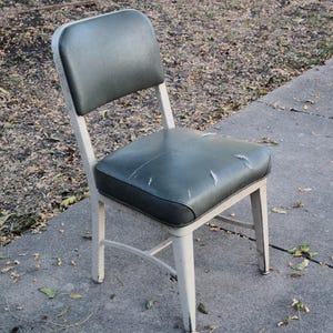 May include: A vintage chair with a dark green seat and backrest. The seat shows signs of wear. The chair has a white metal frame. It is positioned on a concrete surface.