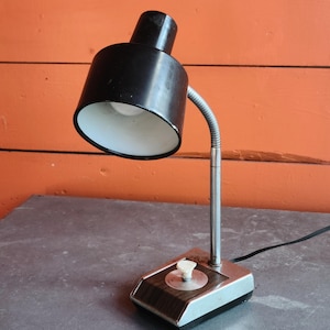May include: A vintage black desk lamp with a flexible, silver-colored neck. The lamp has a white interior and a small, round on/off switch. The base is silver and brown. A black power cord extends from the base.