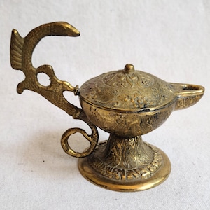 May include: A detailed, antique brass genie lamp with an ornate design. The lamp features a decorative handle, a detailed lid, and a round base. The brass has a warm, aged patina, and the lamp is likely a decorative item.